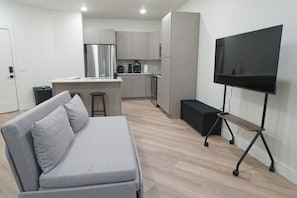 A modern living room with a grey sofa, a flat-screen TV on a stand, a kitchen area with a fridge and coffee maker, and a wooden floor.