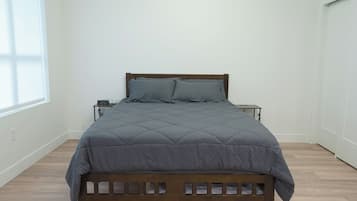 Una cama individual con estructura de madera y colcha gris acolchada en una habitación con paredes blancas y suelo de madera.