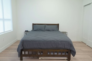 A single bed with a wooden frame and a grey quilted bedspread in a room with white walls and a wooden floor.