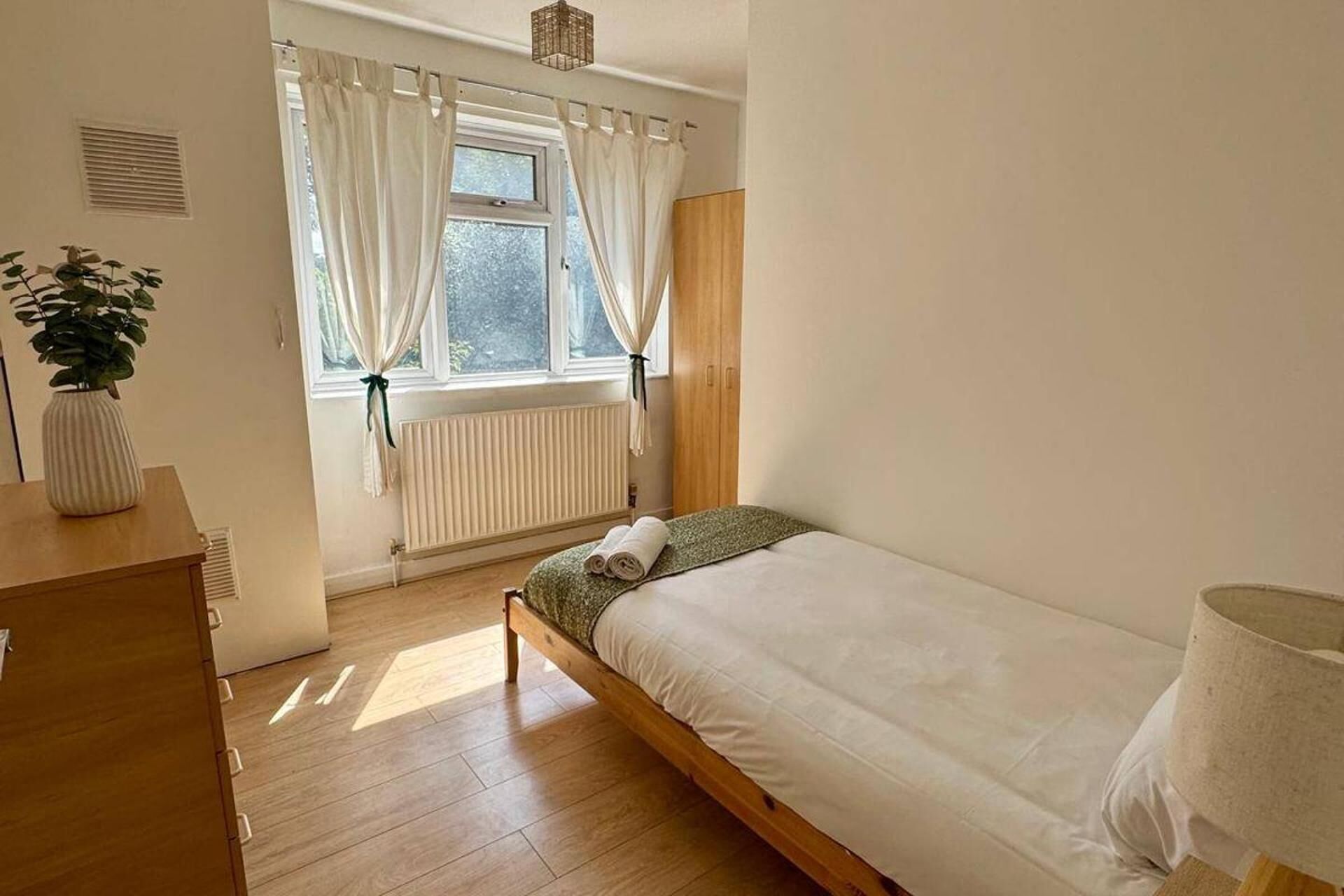 A small, tidy bedroom with a single bed, a bedside table, a radiator, and a window with curtains.