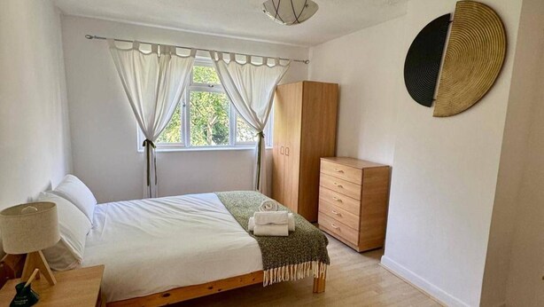 A bedroom with a bed, a wooden wardrobe, a bedside table with a lamp, and a window with curtains. - House Sleeps 8 With Balcony (London)