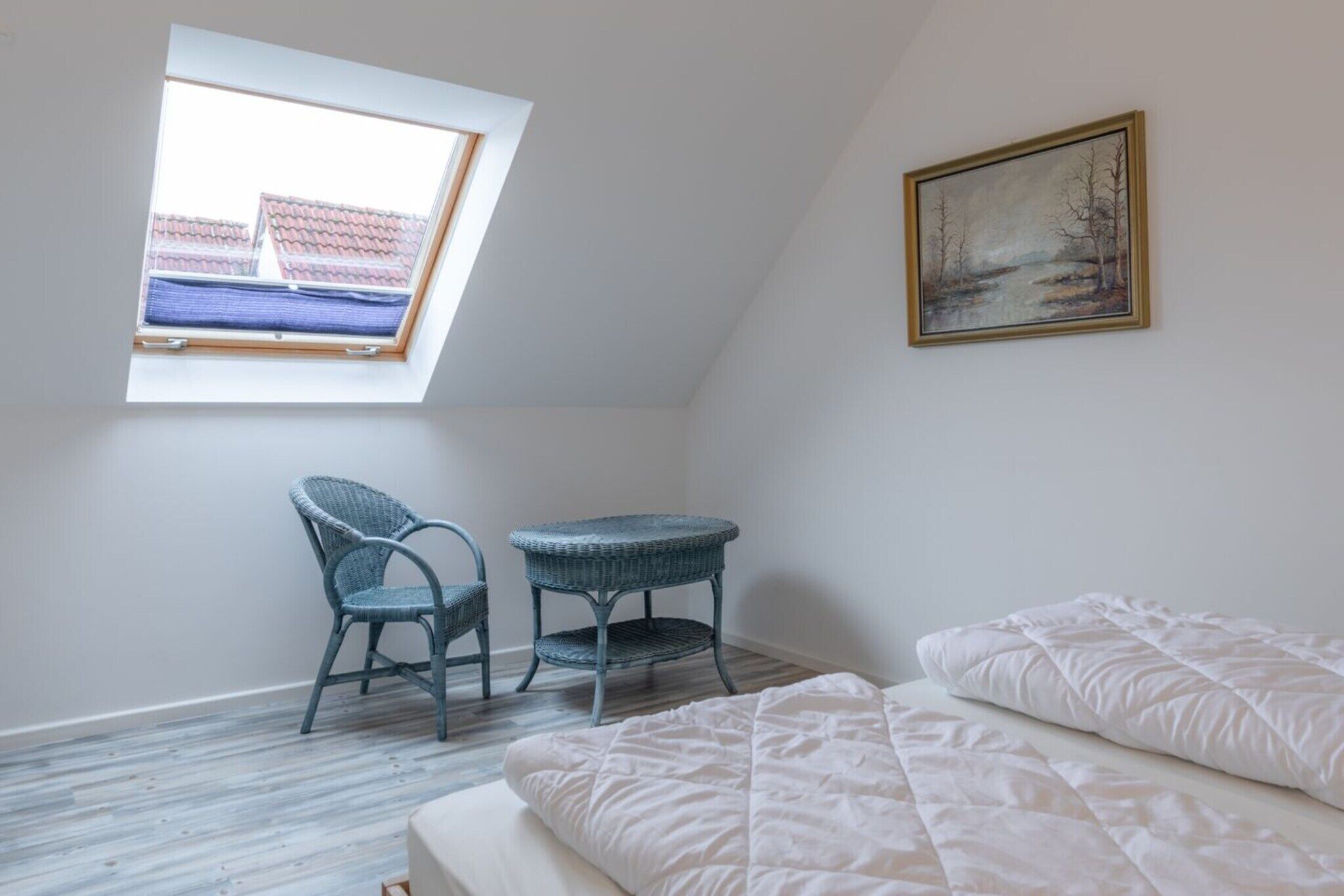 Ein Zimmer mit schrägverdeckter Decke, einem Fenster mit Blick auf Gebäude, einem Sessel, einem kleinen Tisch und einem Bett mit weißer Bettwäsche.
