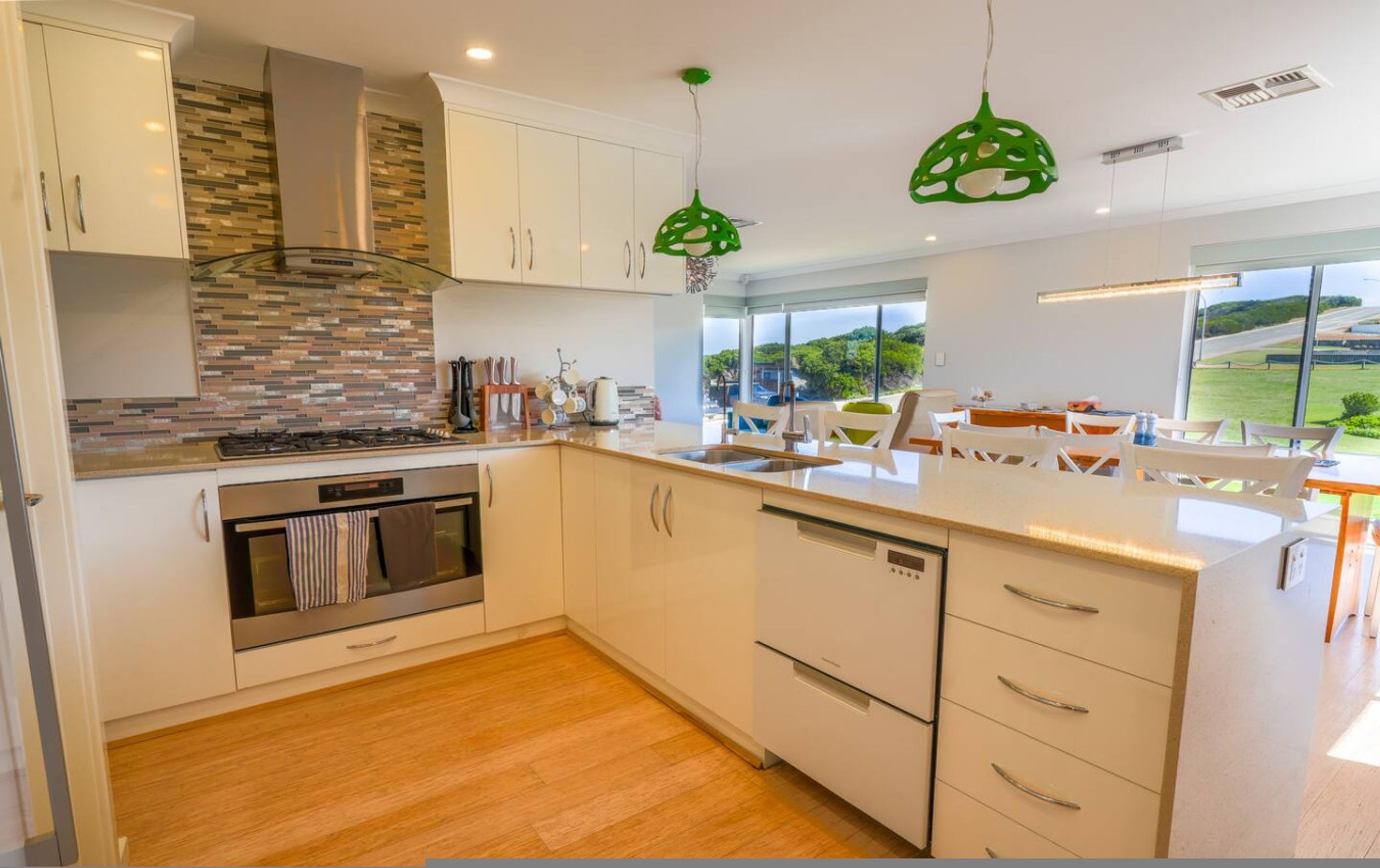 Una cocina moderna con armarios blancos, horno empotrado y lavavajillas. Hay lámparas colgantes verdes y vistas al exterior a través de amplias ventanas.