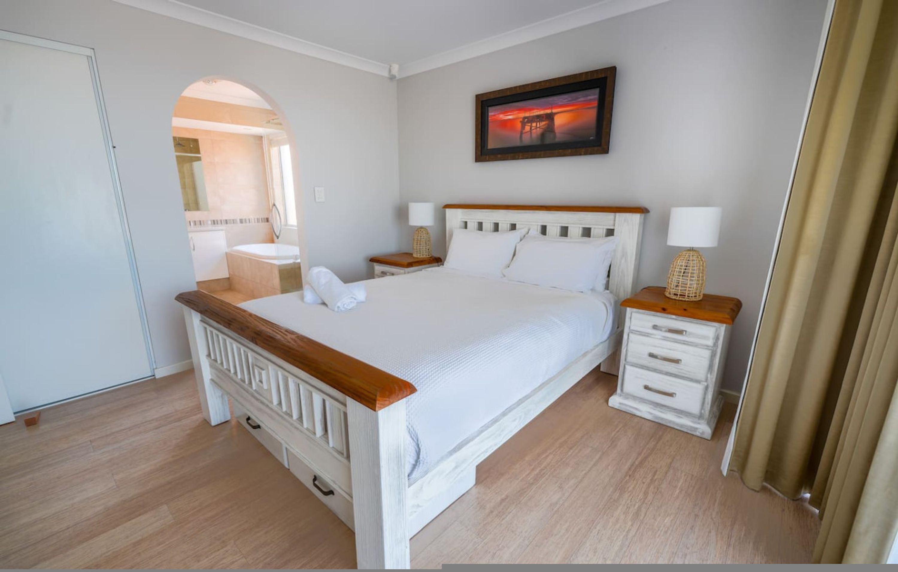 Un dormitorio con cama de madera, mesitas de noche blancas, una lámpara de noche y un cuadro en la pared.