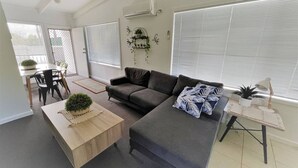 A modern living room with a grey sofa, a wooden coffee table, and a small side table with a lamp. There is a dining area with a table and chairs, and a large window with blinds. - Esperance Little Units 1 (Esperance)