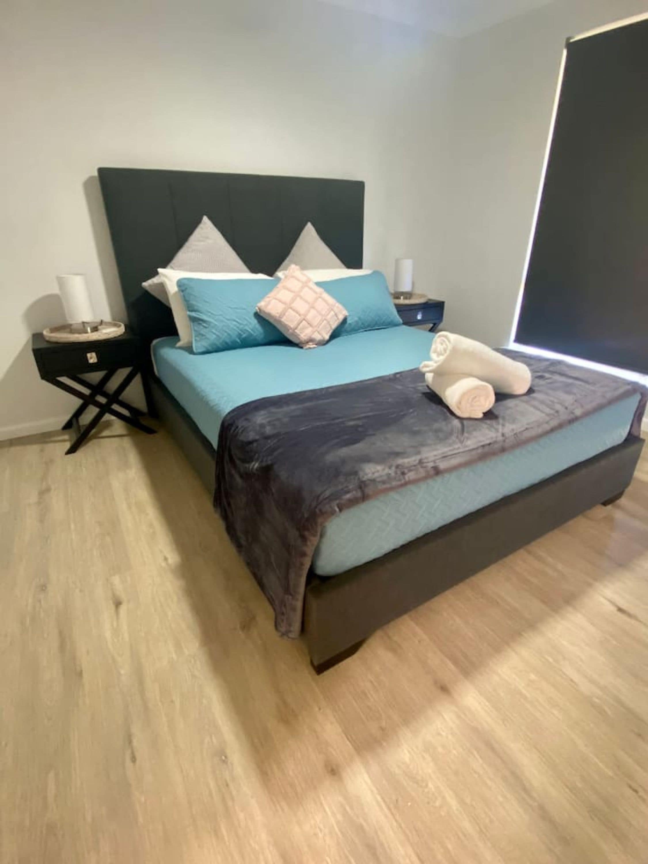 A bed with a dark headboard, a teal bedspread, and a rolled towel on the footboard.