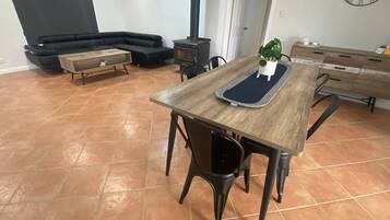 A dining area with a wooden table, chairs, and a bench. There is a black sofa and a small table with a plant in the background.