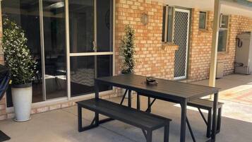 An outdoor patio area with a picnic table and benches, a potted plant, and a building with a brick facade and large windows.