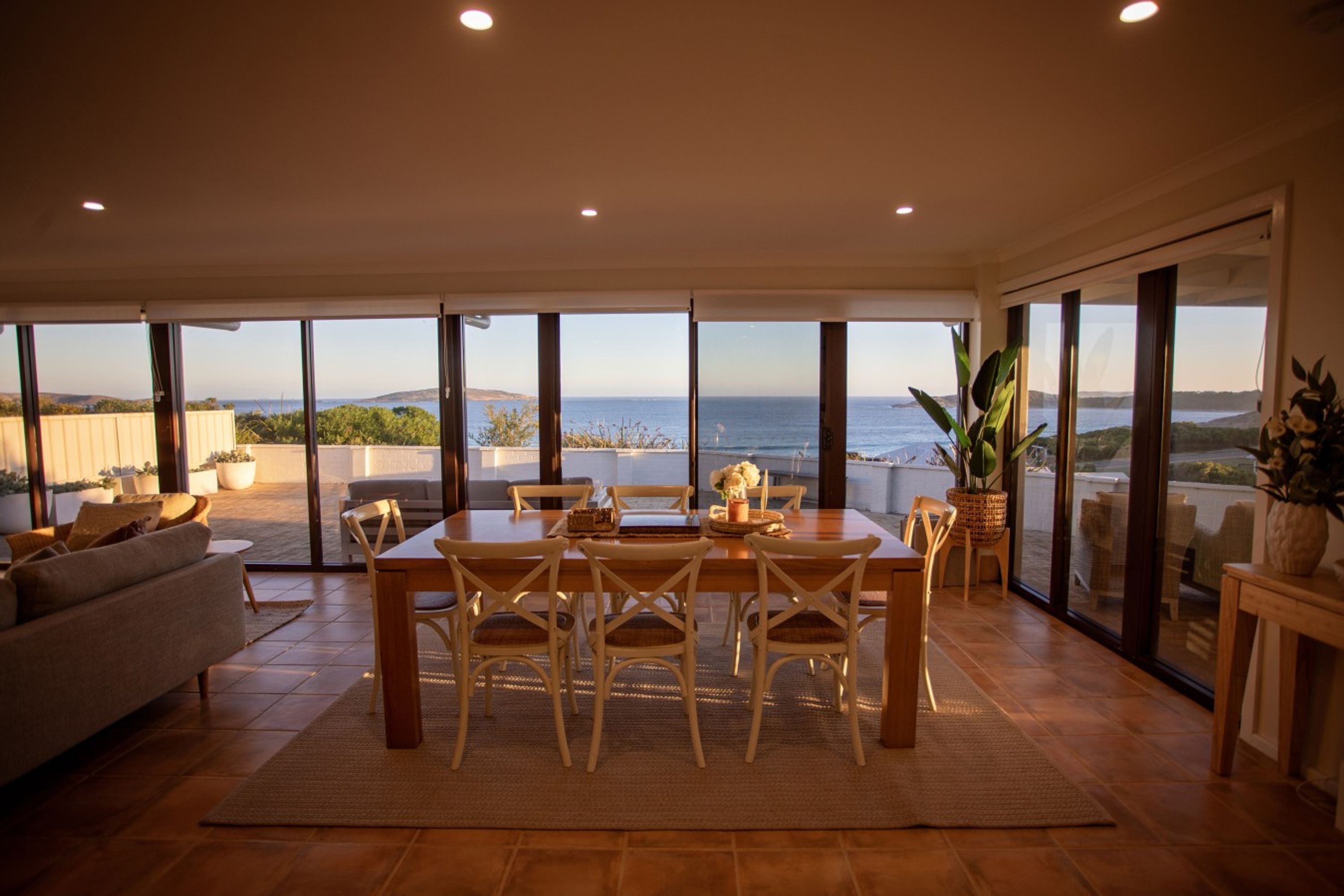 A dining area with a table and chairs, large windows with a view of the sea, and a sofa in the foreground.