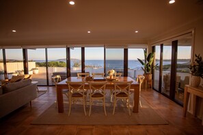 A dining area with a table and chairs, large windows with a view of the sea, and a sofa in the foreground. - West Beach Holiday Home (Esperance)