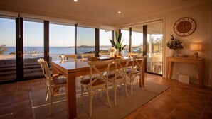 A dining area with a table and chairs, a clock on the wall, and a view of the sea. - West Beach Holiday Home (Esperance)