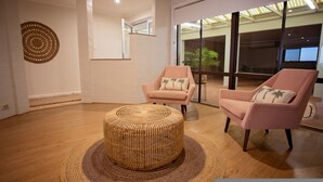 A modern living room with two pink armchairs, a woven ottoman, and a large window offering a view of the outdoors. - West Beach Holiday Home (Esperance)
