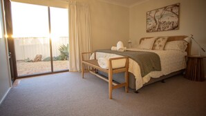A bedroom with a bed, a chair, a bedside table, a lamp, and a sliding glass door leading to an outdoor area. - West Beach Holiday Home (Esperance)