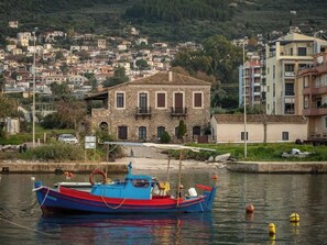 A boat docked near a stone building with a tiled roof, surrounded by calm water and a hillside town in the background. - Charming Beachfront Haven, Steps from the Beach (Kiparissia)
