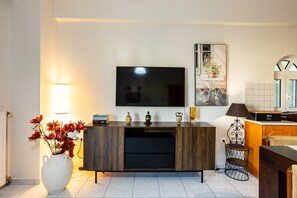 Un salon moderne doté d’un téléviseur à écran plat, d’un meuble de rangement en bois, d’un vase blanc orné de fleurs et d’un coin cuisine équipé d’un four à micro-ondes et d’un grille-pain.