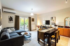 Ein modernes Wohnzimmer mit einer Couch, einem Couchtisch und einem Essbereich mit Stühlen. An der Wand ist ein Fernseher angebracht, es gibt eine Küchenzeile mit Herd und eine Glastür, die auf einen Balkon führt.
