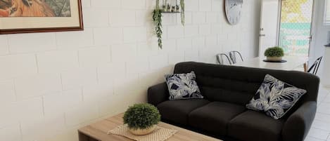 A living room with a dark sofa, a wooden coffee table, a framed picture, a wall clock, and a small plant shelf.