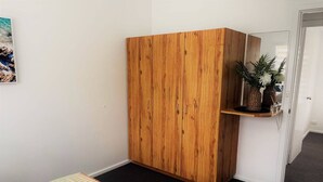 A room with a wooden wardrobe, a bed with striped bedding, and a small shelf with plants. - Esperance Little Units 4 (Esperance)