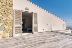 A modern building with a stone facade, a balcony with wooden flooring, and a bedroom with a bed and a blue bedspread. - Villa Skyline: Design, Pool, Ocean View by Sicily Home Solutions (Castellammare del Golfo)