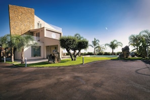 A modern building with a stone facade, a paved driveway, and a well-maintained lawn with palm trees. - Villa Skyline: Design, Pool, Ocean View by Sicily Home Solutions (Castellammare del Golfo)