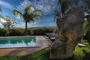 A swimming pool with lounge chairs, surrounded by greenery and a large rock. - Villa Skyline: Design, Pool, Ocean View by Sicily Home Solutions (Castellammare del Golfo)