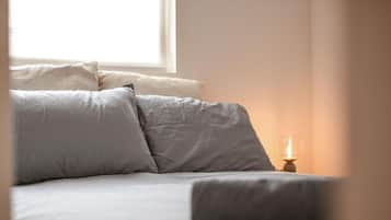 Uma cama com roupa de cama cinza, uma mesa de cabeceira com uma vela acesa e uma janela ao fundo.