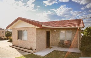 Una casa de un solo piso con techo de tejas rojas, una puerta principal y un pequeño porche con dos sillas.