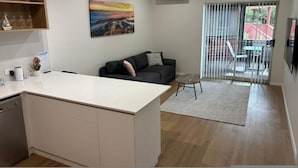A modern kitchen with a white countertop and cabinets, a dining area with a black sofa and a small table, and a large window with blinds. - Seabreeze Esperance in West Beach (Esperance)
