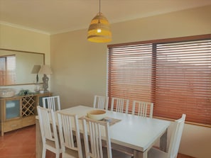 A dining area with a white table and chairs, a hanging light fixture, a window with blinds, and a wooden cabinet with a mirror and lamp. - The Oceanaire in West Beach (Esperance)