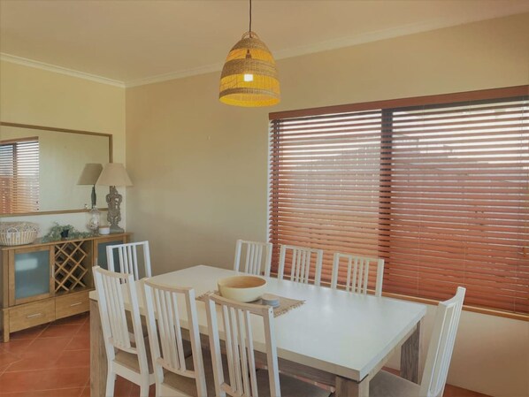 A dining area with a white table and chairs, a hanging light fixture, a window with blinds, and a wooden cabinet with a mirror and lamp.