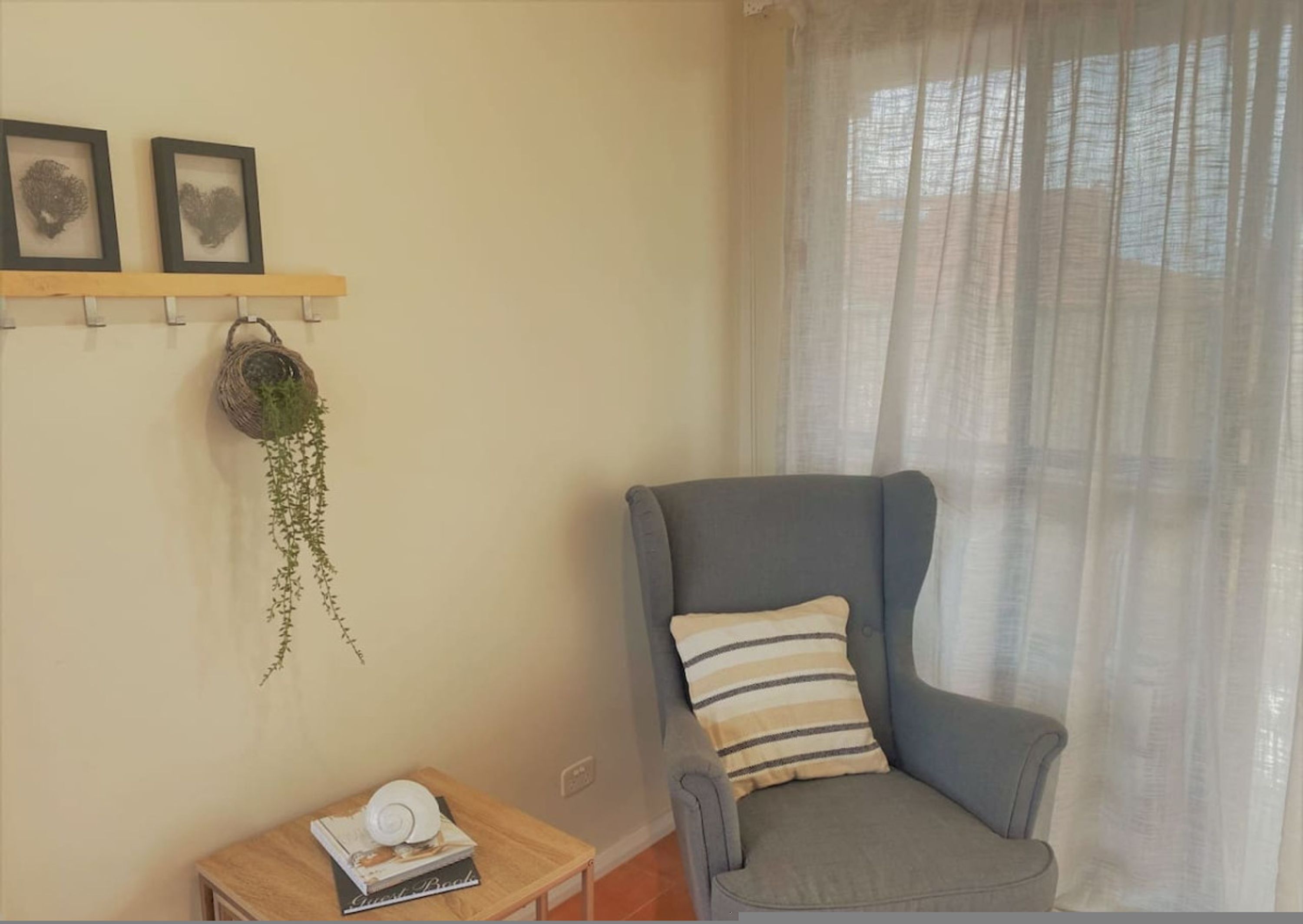 A room with a grey armchair, a small wooden table with a book, a hanging plant, and two framed pictures on the wall.