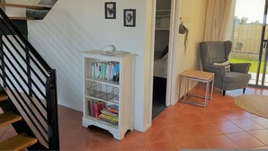 A staircase leading to a landing, a white bookshelf with books, a grey armchair, and a view of a garden through a sliding door.