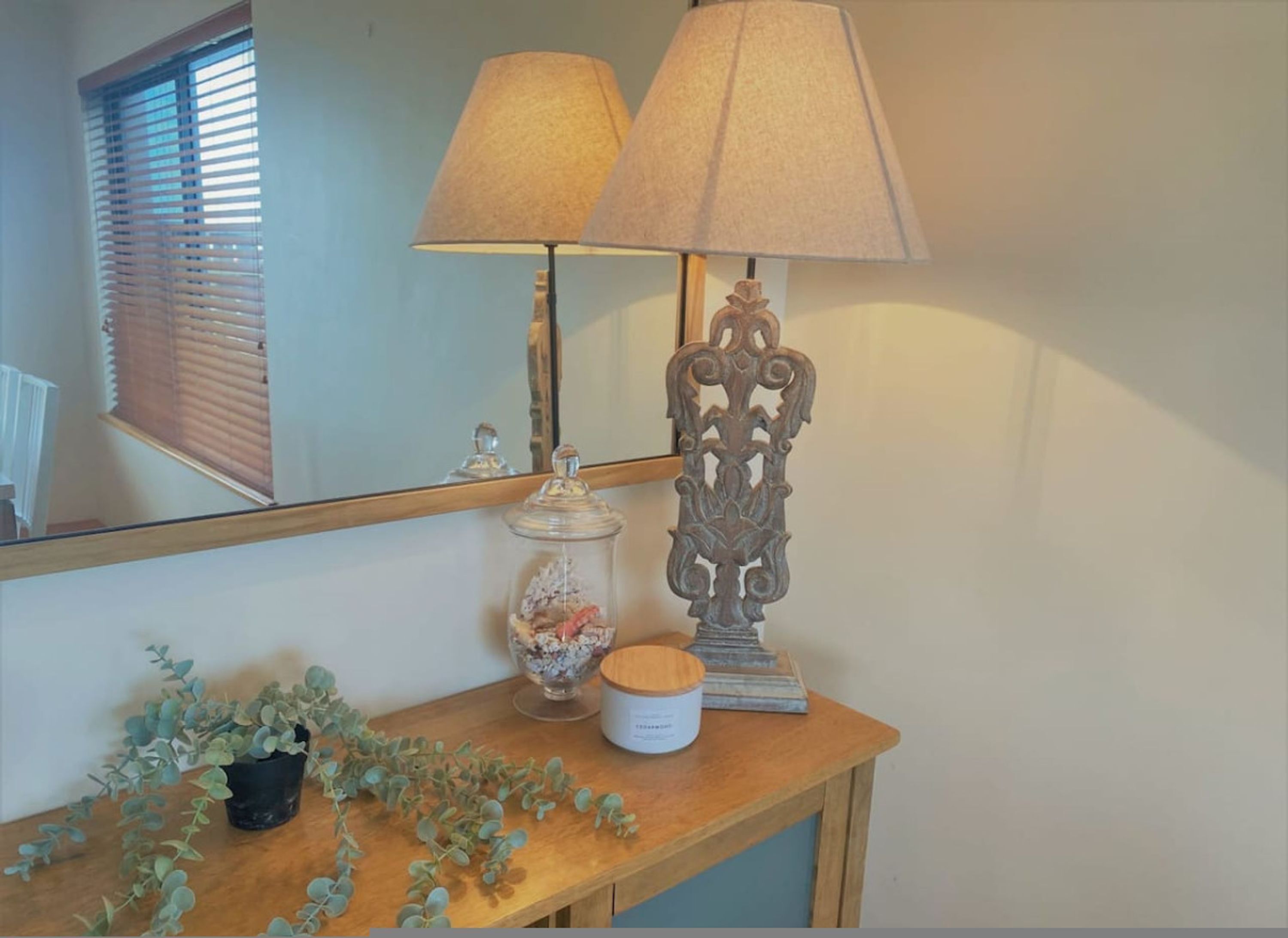 Una consola de madera con una lámpara, un espejo decorativo, un tarro de cristal con flores y una pequeña planta en maceta.