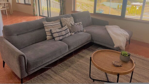 A grey sofa with pillows, a woven coffee table, and a view of a residential area through large windows.