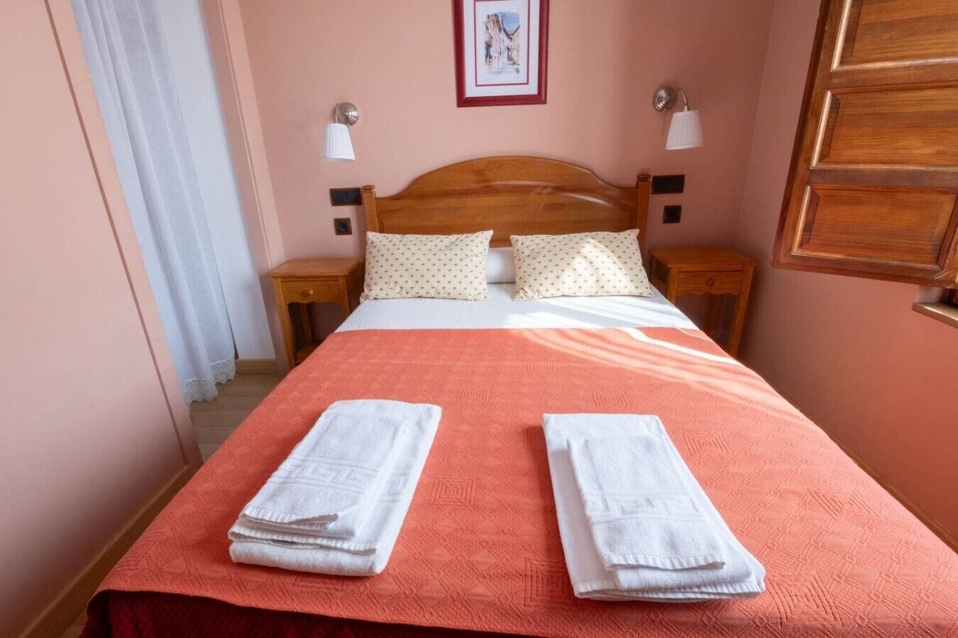 Een netjes opgemaakt bed met twee kussens, een rode spreideken en twee witte handdoeken die erover gevouwen zijn.
