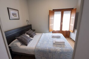 A bedroom with a bed, pillows, a bedside table, a lamp, a window with wooden shutters, and a framed picture on the wall.