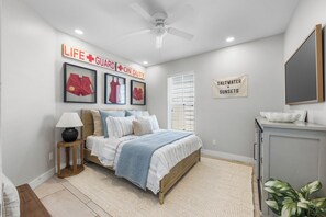 Ein Schlafzimmer mit einem Bett, einem Nachttisch, einer Lampe, einem Deckenventilator, gerahmten Bildern an der Wand und einem Schild mit der Aufschrift 'LEBENSRETTUNGSDIENST EINSATZBEREIT'.