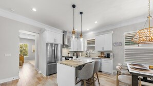 A modern kitchen with a central island, stainless steel appliances, pendant lighting, and a view of the adjacent living area. - Sunny 3BR Getaway | Good Morning Rentals (Jacksonville Beach)