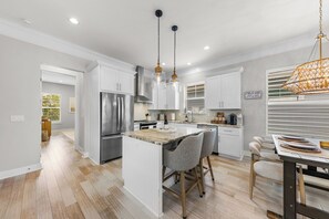 A modern kitchen with a central island, stainless steel appliances, pendant lighting, and a view of the adjacent living area. - Sunny 3BR Getaway | Good Morning Rentals (Jacksonville Beach)