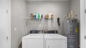 A laundry area with two washing machines, a water heater, and a shelf with cleaning supplies. - Sunny 3BR Getaway | Good Morning Rentals (Jacksonville Beach)