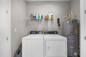 A laundry area with two washing machines, a water heater, and a shelf with cleaning supplies. - Sunny 3BR Getaway | Good Morning Rentals (Jacksonville Beach)