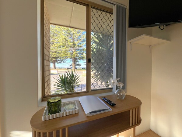 A room with a wooden desk, a window with a view of trees and a road, a TV mounted on the wall, and a small plant on the desk.