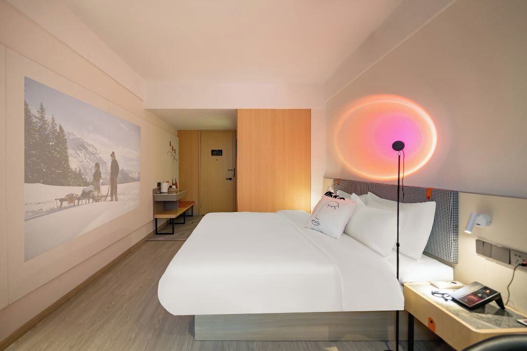 A hotel room with a large bed, a desk, a chair, a nightstand, and a wall-mounted lamp.