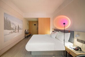 A hotel room with a large bed, a desk, a chair, a nightstand, and a wall-mounted lamp. - Sushi Light Luxury Hotel Xingzhuang (Shanghai)