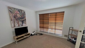 A room with a flat-screen TV on a stand, a painting on the wall, a window with blinds, and a door with a shelf. - The Willow in West Beach (Esperance)