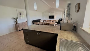 A modern kitchen with a central island, dining area with chairs, and a TV mounted on the wall. - The Willow in West Beach (Esperance)