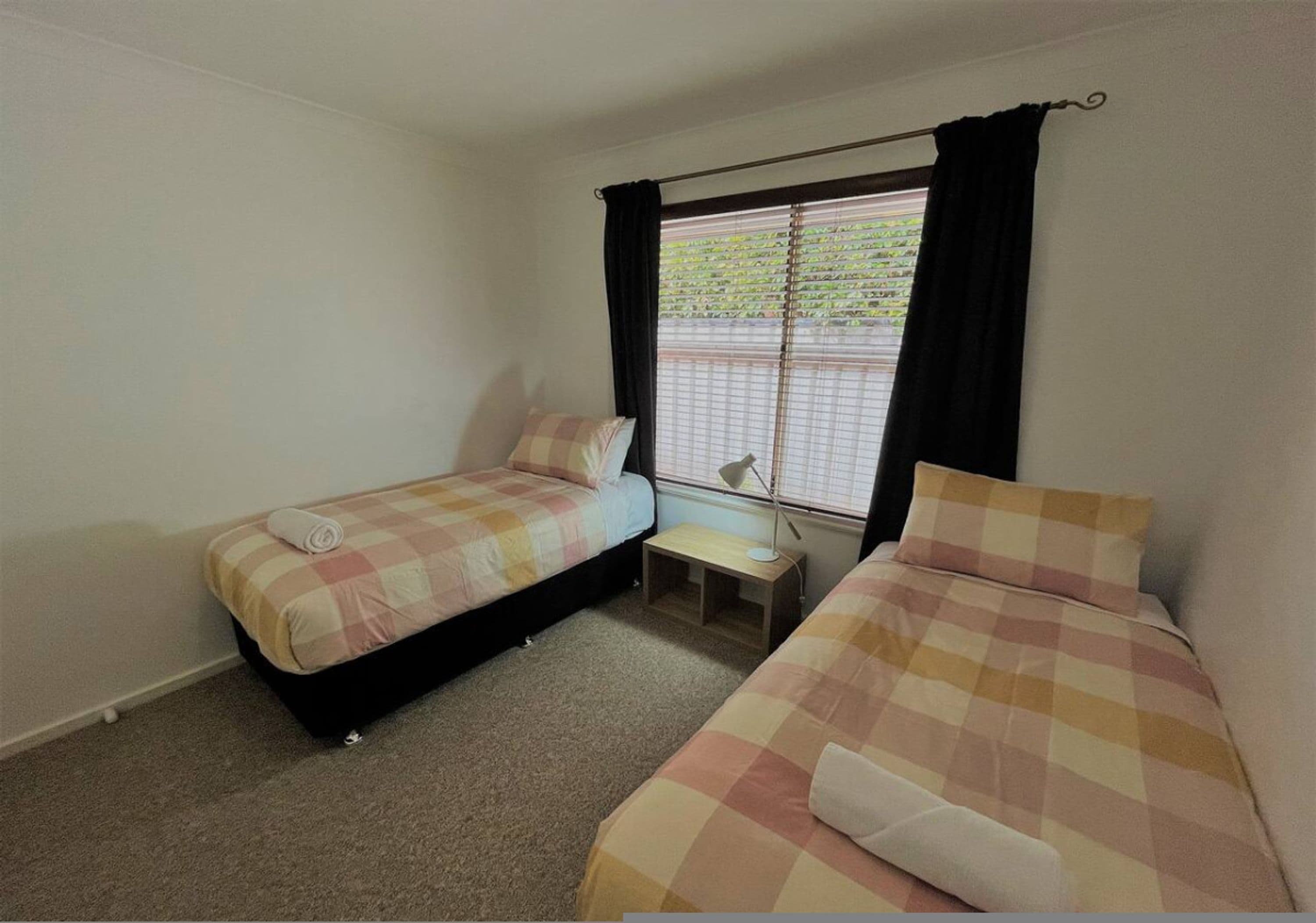 A small, single-room accommodation with two beds, a window with blinds, a bedside table, and a lamp.