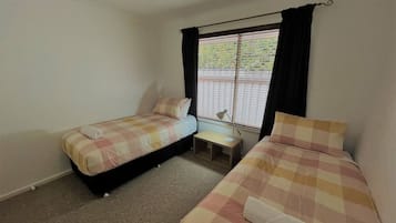 Un alojamiento pequeño de una sola habitación con dos camas, una ventana con persianas, una mesita de noche y una lámpara.