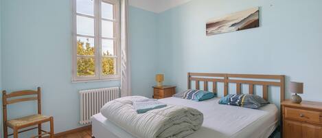 A bedroom with a bed, a chair, a nightstand, a lamp, and a window.