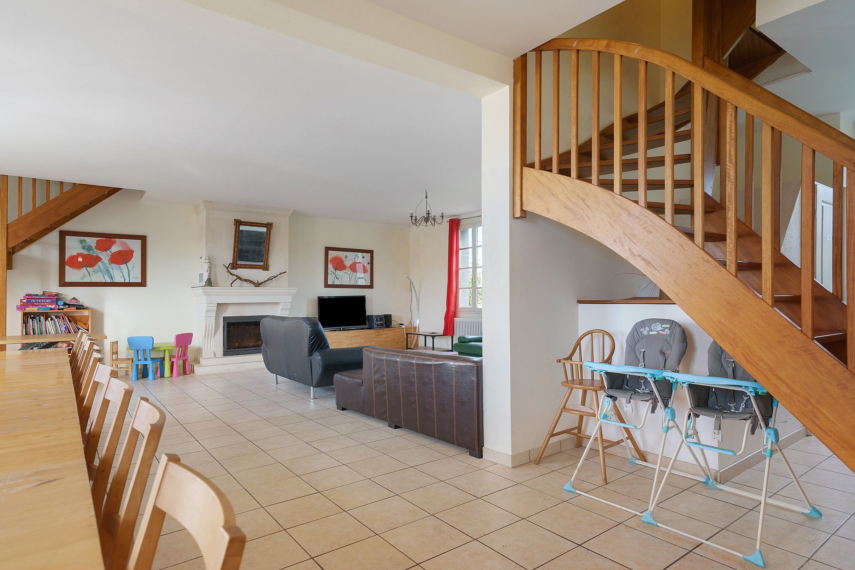 A spacious living room with a fireplace, a dining area with chairs, a staircase with wooden railings, and a baby play area with a changing station.